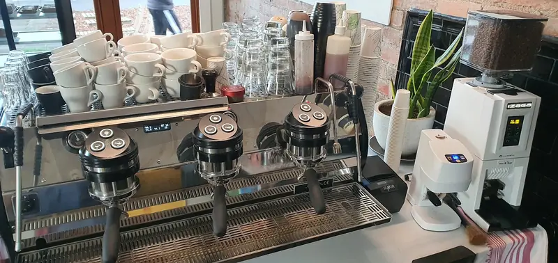 Mitte's espresso bar: three-group machine, stacked white cups and glasses, grinders and a snake plant