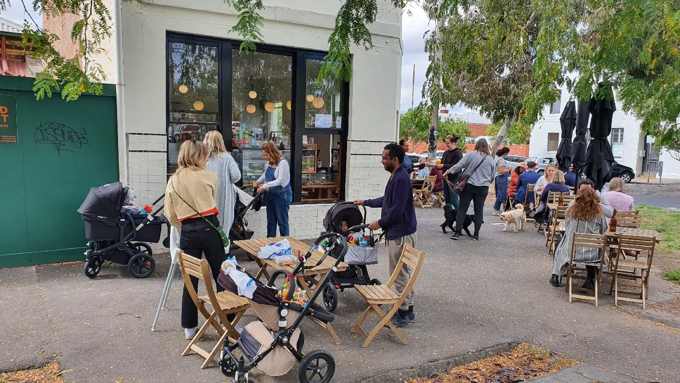 Mitte's white-tiled shopfront on a busy weekend, with families, prams and dogs at the timber footpath tables under the trees