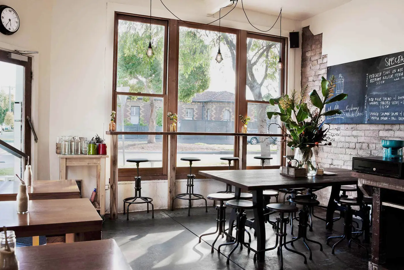 Inside Mitte: tall timber-framed windows onto a leafy street, industrial stools at a long timber table, and a chalkboard of specials on the exposed brick wall